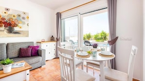 A bright living room with a gray sofa, a dining table with white chairs, and a large window with curtains.