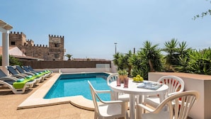A pool area with lounge chairs and a table, a castle-like building in the background, and tropical plants.