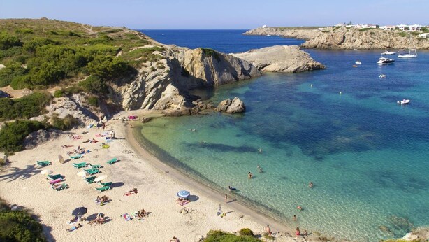 Una playa costera con aguas cristalinas, afloramientos rocosos y algunos barcos.
