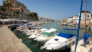 A marina with boats docked, a historic building in the background, and a paved walkway along the water. - Villa Solana by Villa Plus (Ciutadella de Menorca)