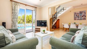 Un salon équipé d’un téléviseur, de canapés verts, d’une table basse et d’un balcon donnant sur la piscine.