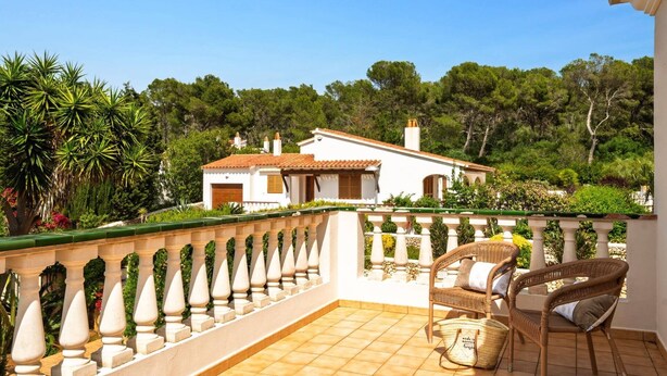 Un balcon avec un sol carrelé, une chaise en osier et un bâtiment blanc en arrière-plan.