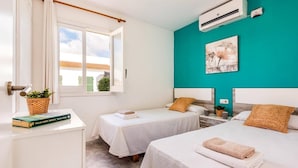 A hotel room with two beds, a window with curtains, a wall-mounted air conditioner, and a teal accent wall. - Villa Pomelo by Villa Plus (Ciutadella de Menorca)