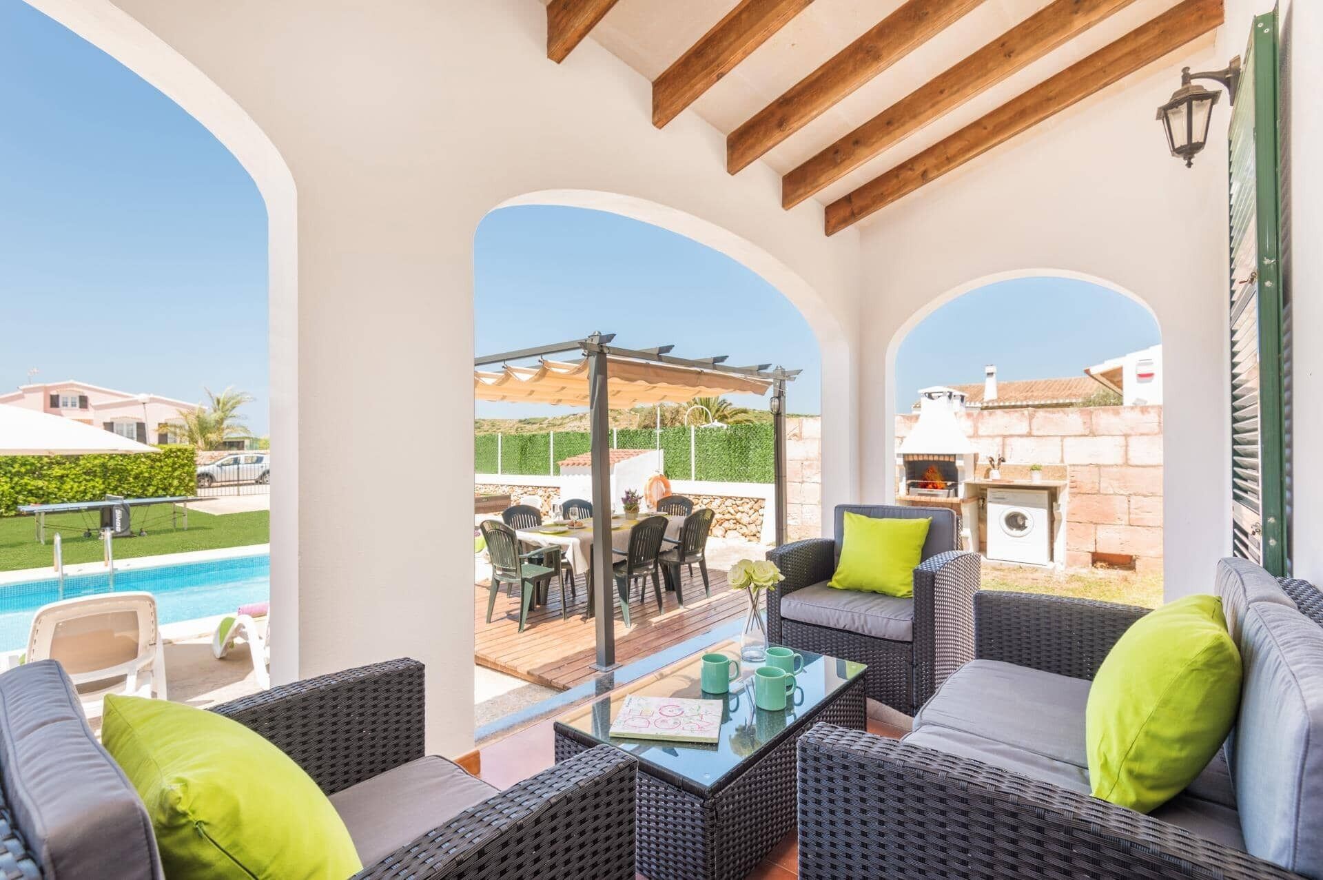 A covered outdoor seating area with wicker furniture and a glass table, overlooking a pool and a garden.