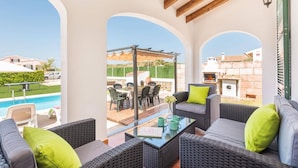 A covered outdoor seating area with wicker furniture and a glass table, overlooking a pool and a garden. - Villa Pomelo by Villa Plus (Ciutadella de Menorca)