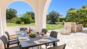 Un área de comedor al aire libre con una mesa dispuesta para cuatro personas, rodeada de sillas y un hogar de piedra al fondo.