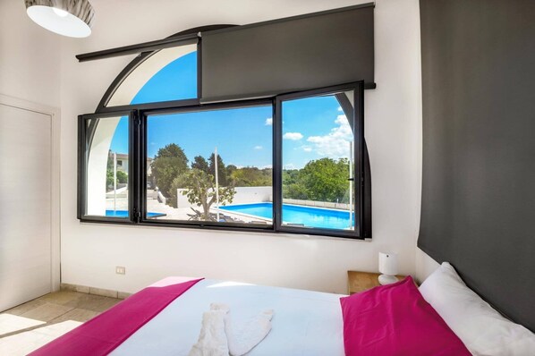 Ein Schlafzimmer mit großem Fenster mit Blick auf einen Pool und Grünflächen.