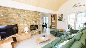 Un salon moderne avec un mur en pierre, un téléviseur à écran plat, une cheminée, un canapé vert et un coin repas à l’arrière-plan.
