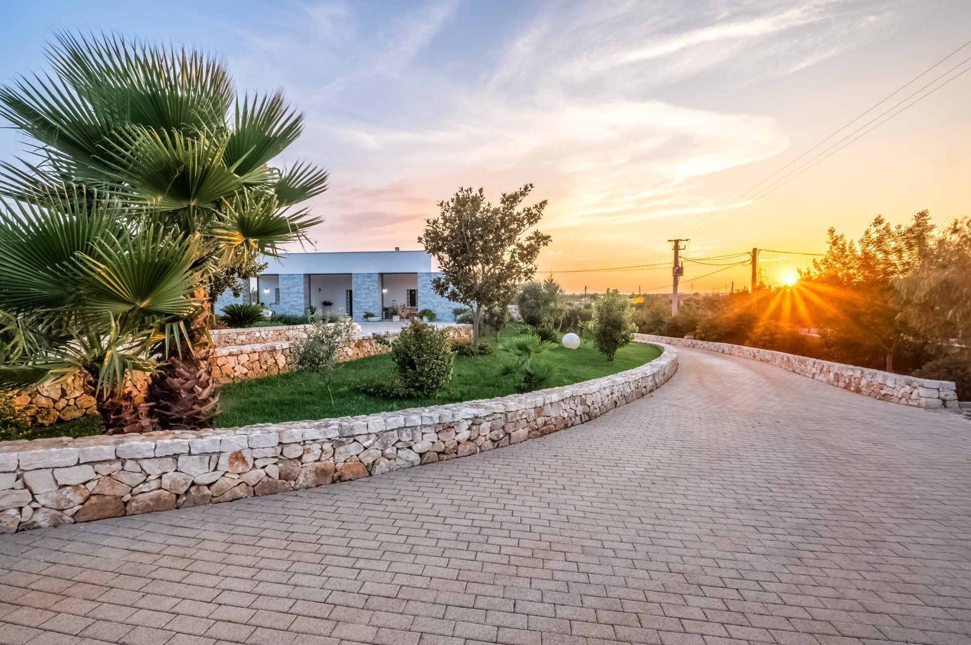 Une maison moderne avec une allée pavée, des murs en pierre et une variété de plantes, notamment des palmiers et des arbustes.