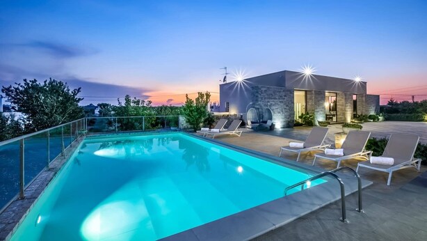 Une piscine avec des chaises longues, un bâtiment en pierre doté de portes vitrées et une vue sur le coucher de soleil.