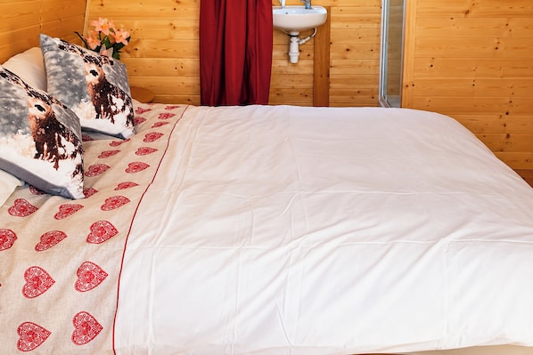 Ein Bett mit weißer Bettwäsche und roten Herzmustern, ein hölzernes Kopfende, ein roter Vorhang und ein weißes Waschbecken in einem Raum mit Holzeinrichtung.
