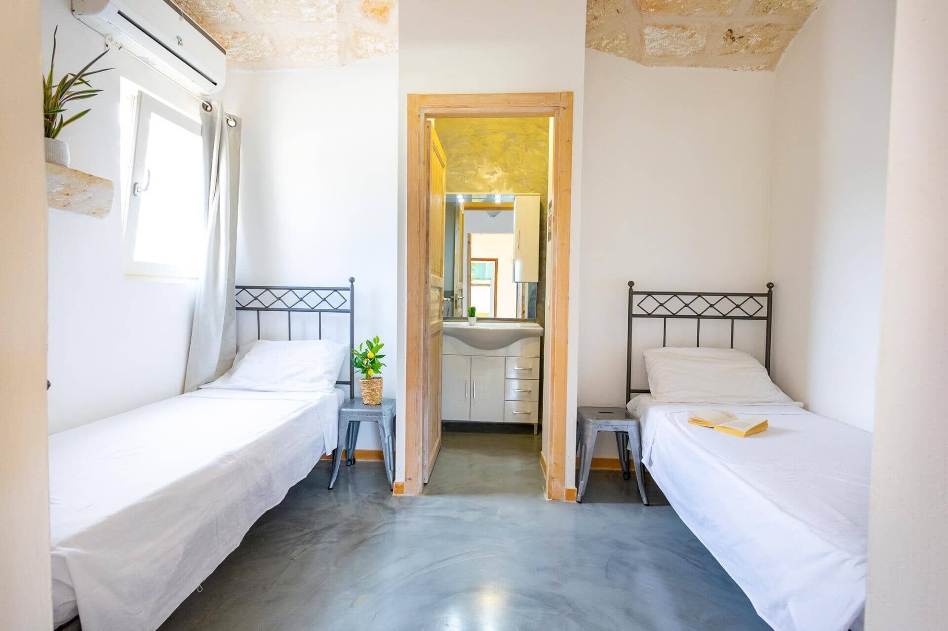 A small, bright room with two single beds, a small table, and a mirror.