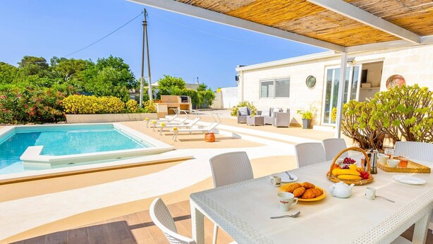Zona de piscina con una mesa lista para el desayuno, sillas de descanso y una casa pequeña al fondo.