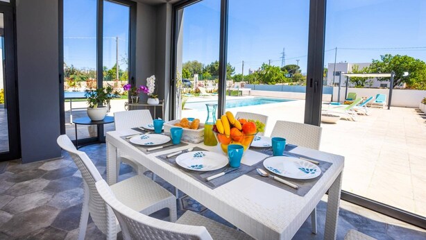 Un coin repas avec une table dressée pour deux personnes, donnant sur une piscine et un espace de détente extérieur.