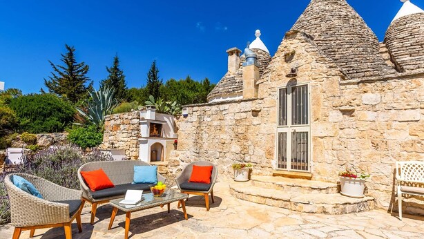 A stone building with a chimney, a window, and a door. There is a glass table with chairs and a potted plant. - Villa Trulli Isabella by Villa Plus (Martina Franca)