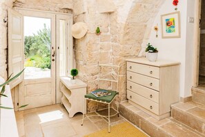 Une pièce avec un mur en pierre, une porte en bois, une petite table avec une plante, une chaise verte et une commode blanche.