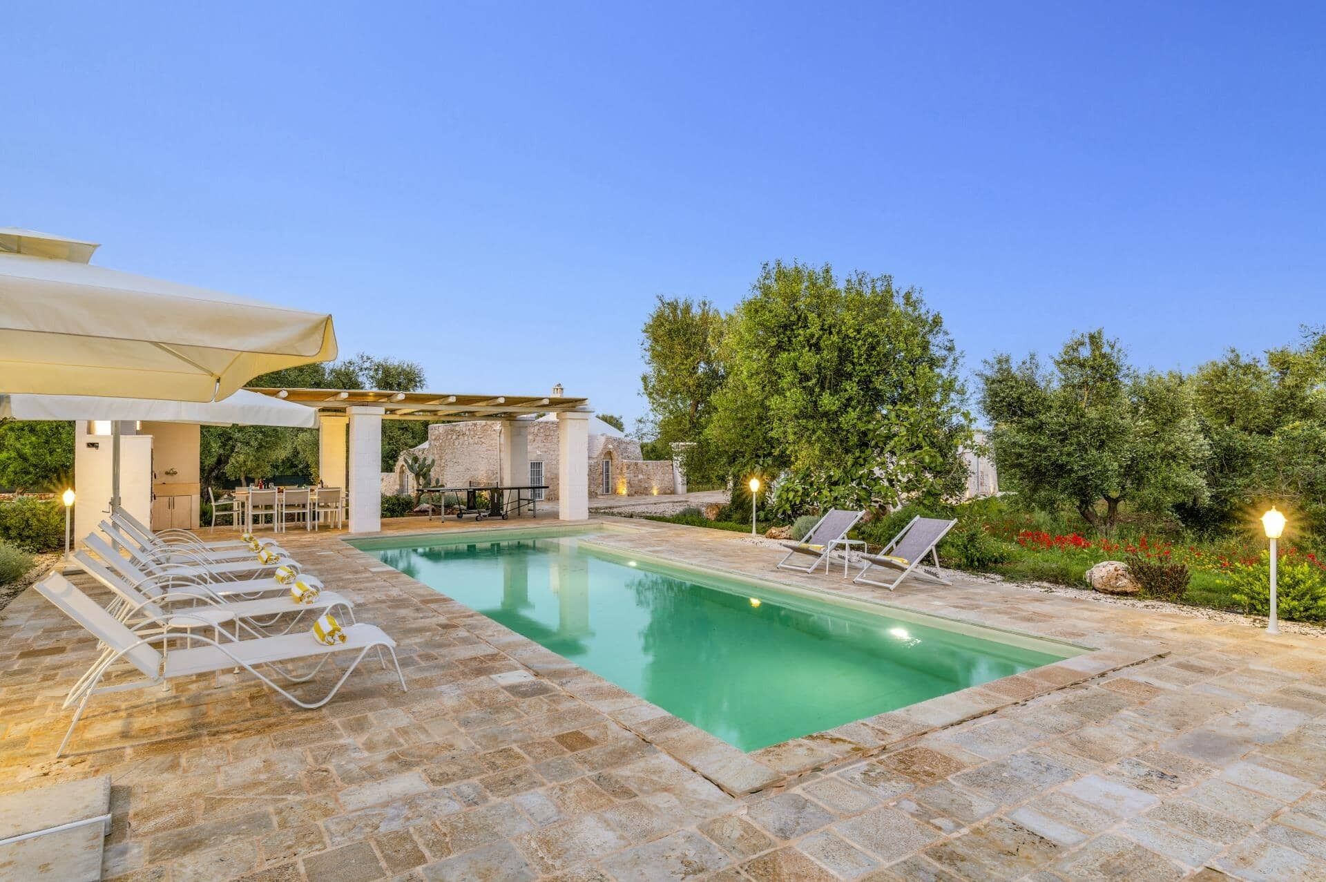 En swimmingpool med solstolar, en pergola och en stenbyggnad i bakgrunden.