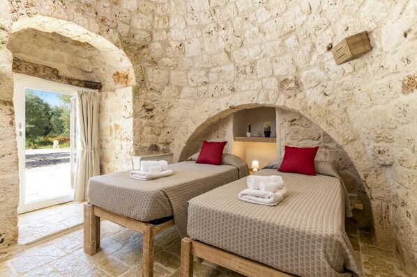 Una habitación con dos camas, paredes de piedra, una mesita y una puerta que da al exterior.