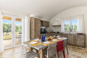A spacious kitchen with a central island, a dining area with a table set for four, and a sliding glass door leading to an outdoor patio. - Villa Trullo Acquaro by Villa Plus (Ostuni)