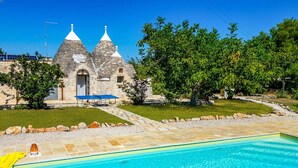 A swimming pool with a stone deck, a stone building with two conical roofs, and a well-maintained lawn with trees. - Villa Trullo Bozalice by Villa Plus (Martina Franca)