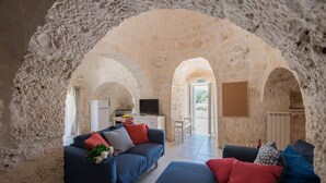 A cozy living room with a stone wall, a sofa set, a television, and a kitchen area visible through an archway. - Villa Trullo Bozalice by Villa Plus (Martina Franca)
