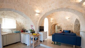 A compact kitchen with a sink, a toaster, and a television. There is a blue sofa and a dining table with chairs. The room has arched ceilings and a view into another room. - Villa Trullo Bozalice by Villa Plus (Martina Franca)