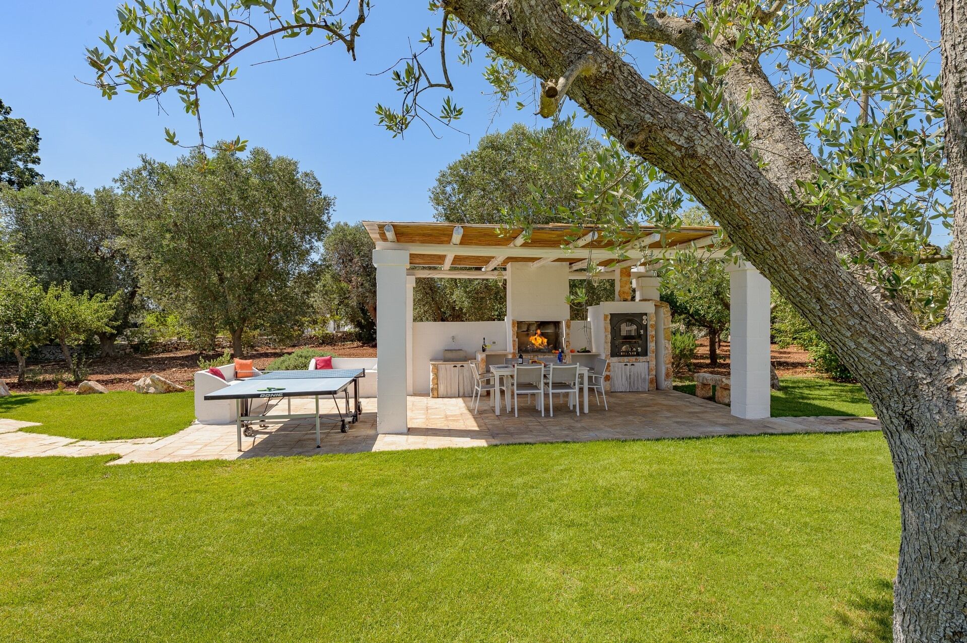 Une terrasse extérieure équipée d’une table de ping-pong, de sièges et d’un espace barbecue aménagé sous une pergola.