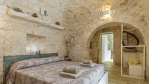 Quarto com paredes de pedra, uma cama, uma estante com objetos decorativos e um cabideiro.
