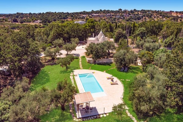 Une grande piscine privée avec une terrasse en pierre, entourée d’une végétation luxuriante et d’un bâtiment en pierre.