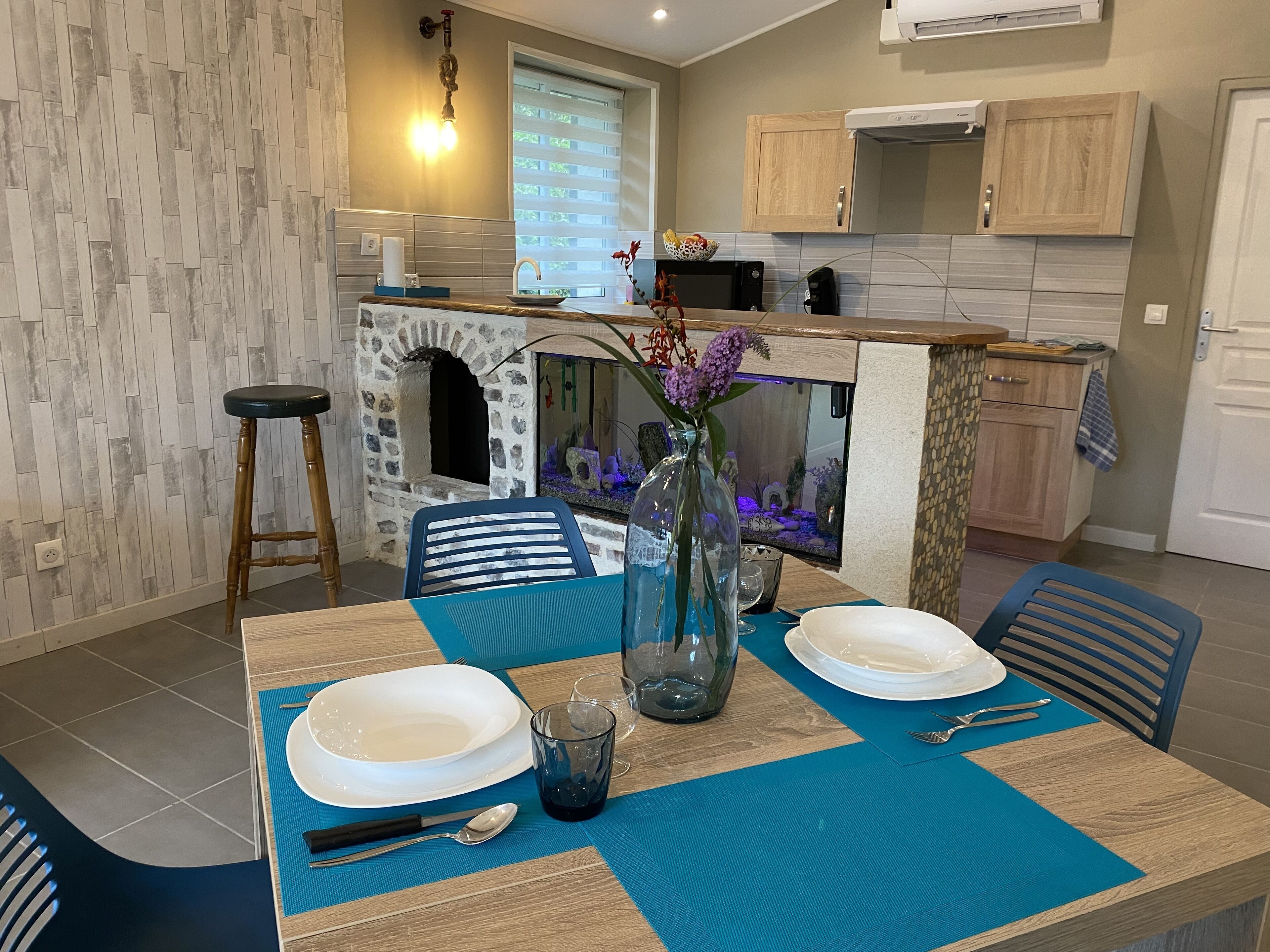 Un comedor con una mesa dispuesta para dos, una chimenea de piedra y una cocina al fondo.
