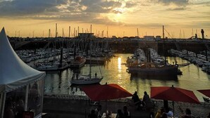A marina with boats docked, a sunset in the background, and people sitting at tables under umbrellas.