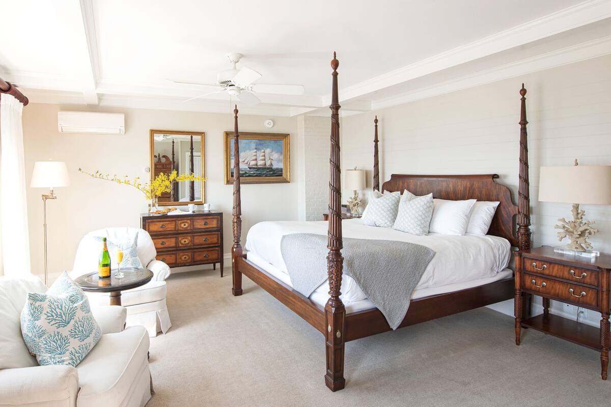 A four-poster bed with a wooden headboard, a nightstand with drawers, a dresser, a chair with a cushion, a floor lamp, and a ceiling fan.