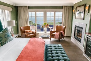 Een hotelkamer met een groot bed, twee stoelen, een klein tafeltje met drankjes, een open haard en uitzicht op zee.