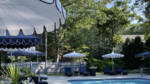 Un área de piscina con sillas de descanso y sombrillas, rodeada de árboles y una valla.