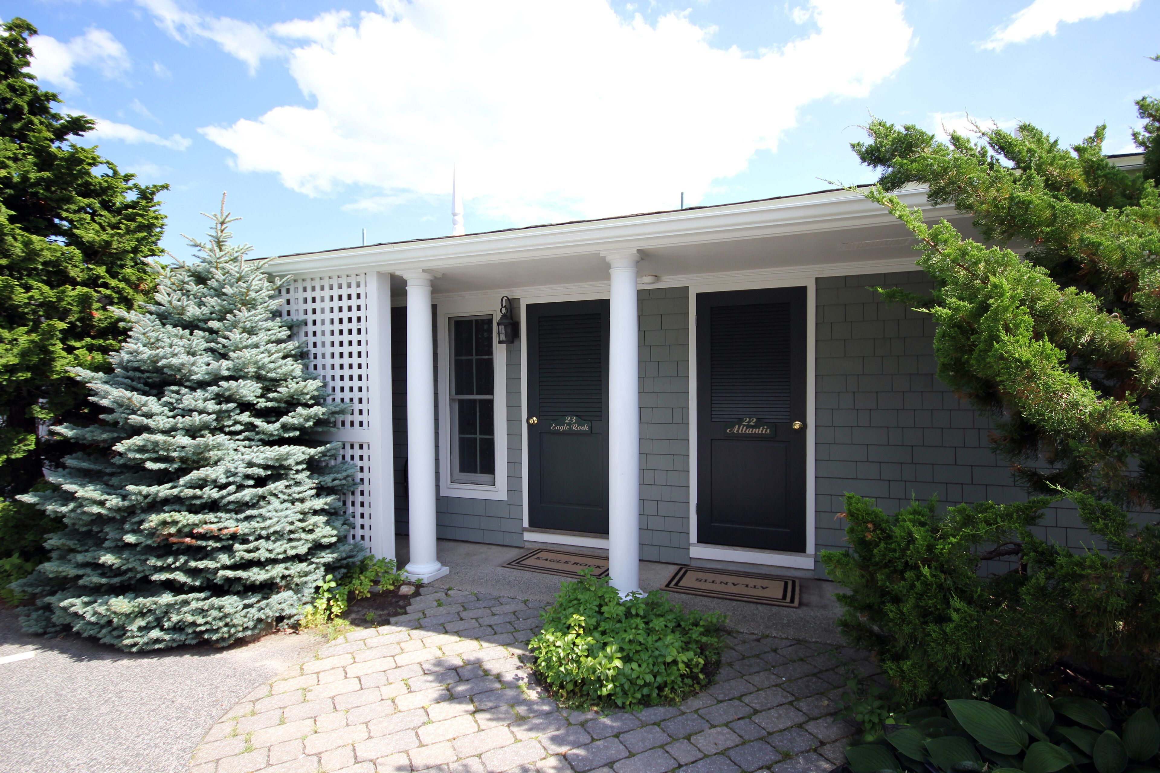 A building with a covered porch, two doors labeled "23" and "22," and a small garden area with a conifer and shrubs.
