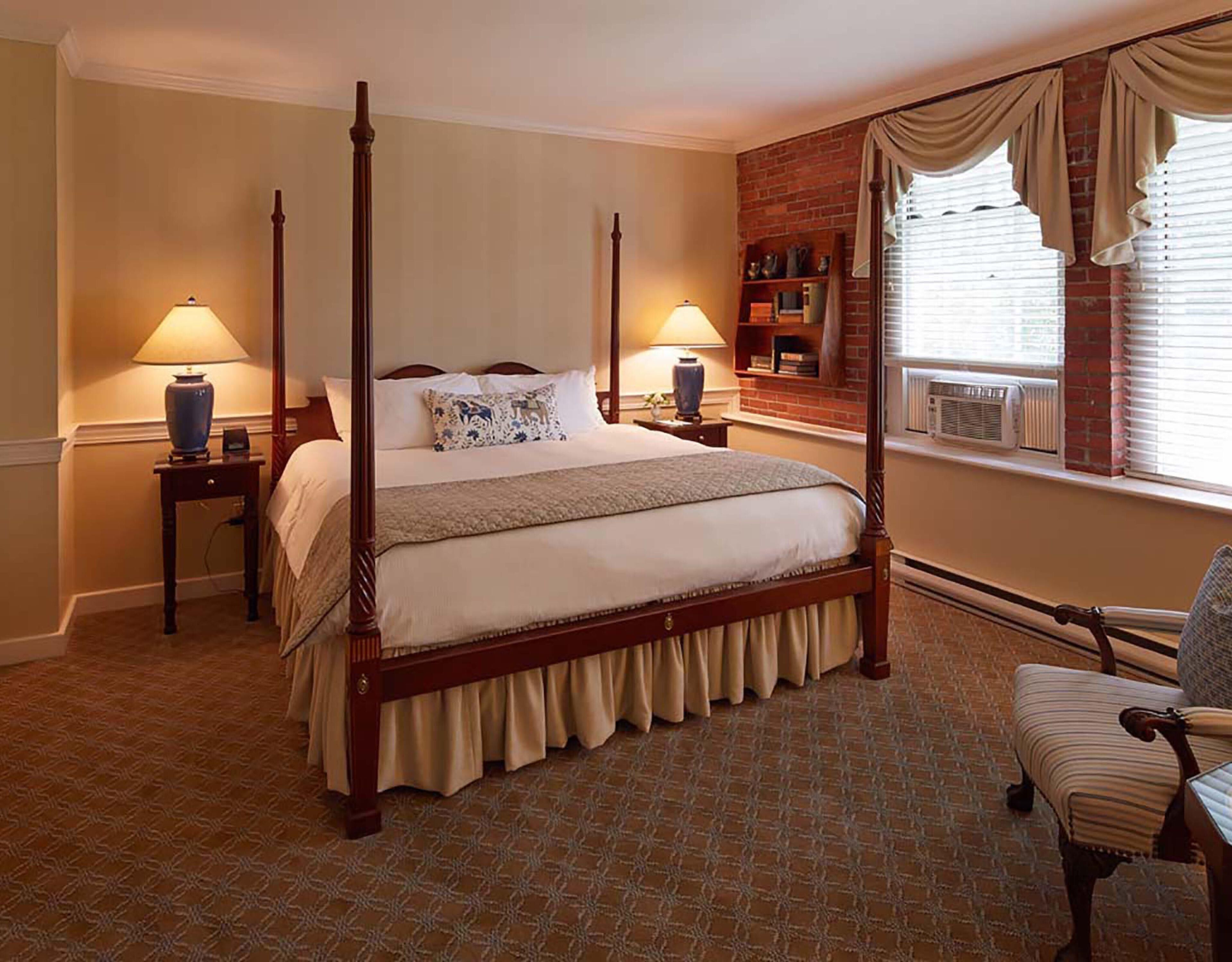 A four-poster bed with a wooden frame, two bedside tables with lamps, a brick wall, a window with curtains, and a patterned carpet.