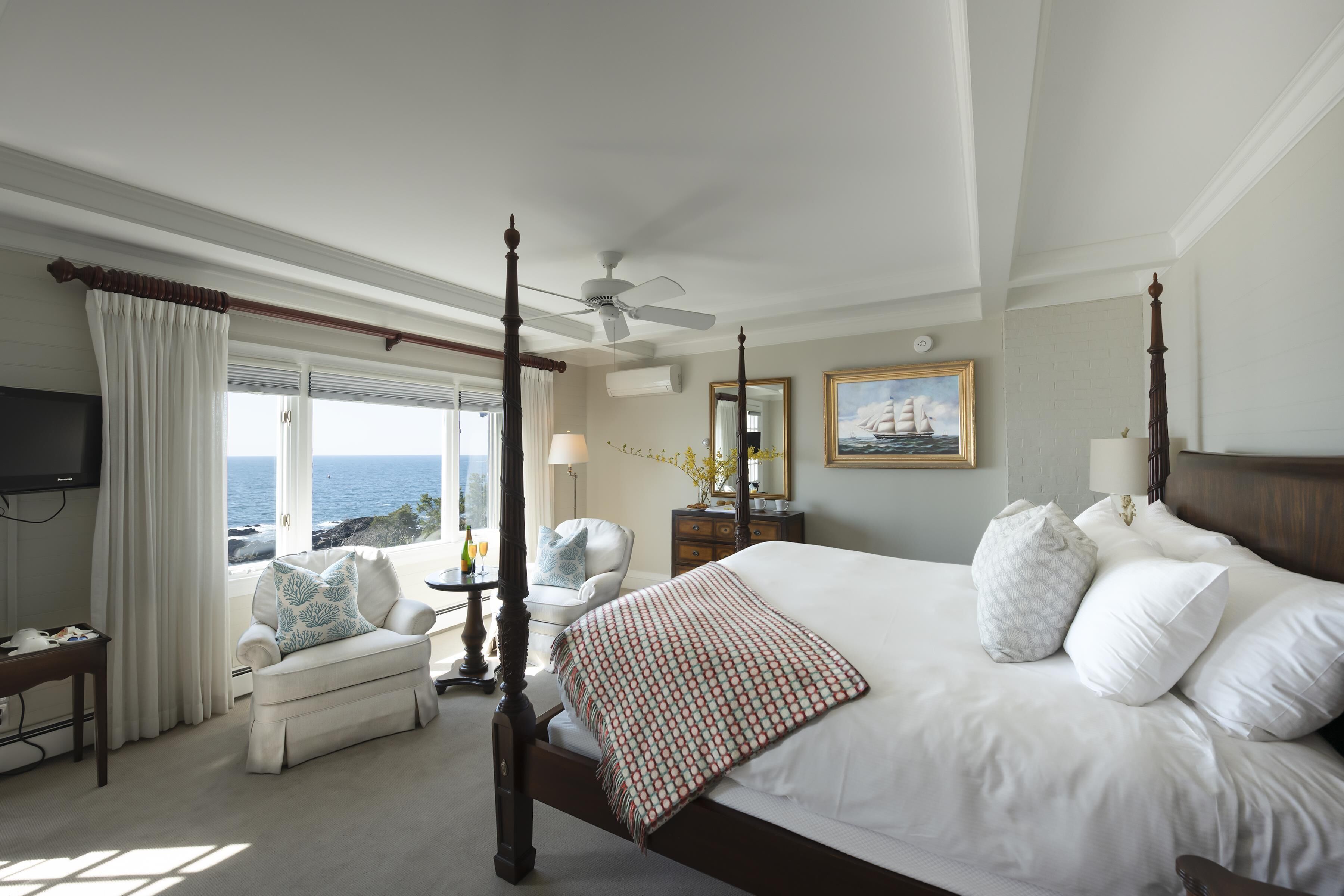 A spacious bedroom with a large bed, a four-poster headboard, a ceiling fan, and a view of the ocean.