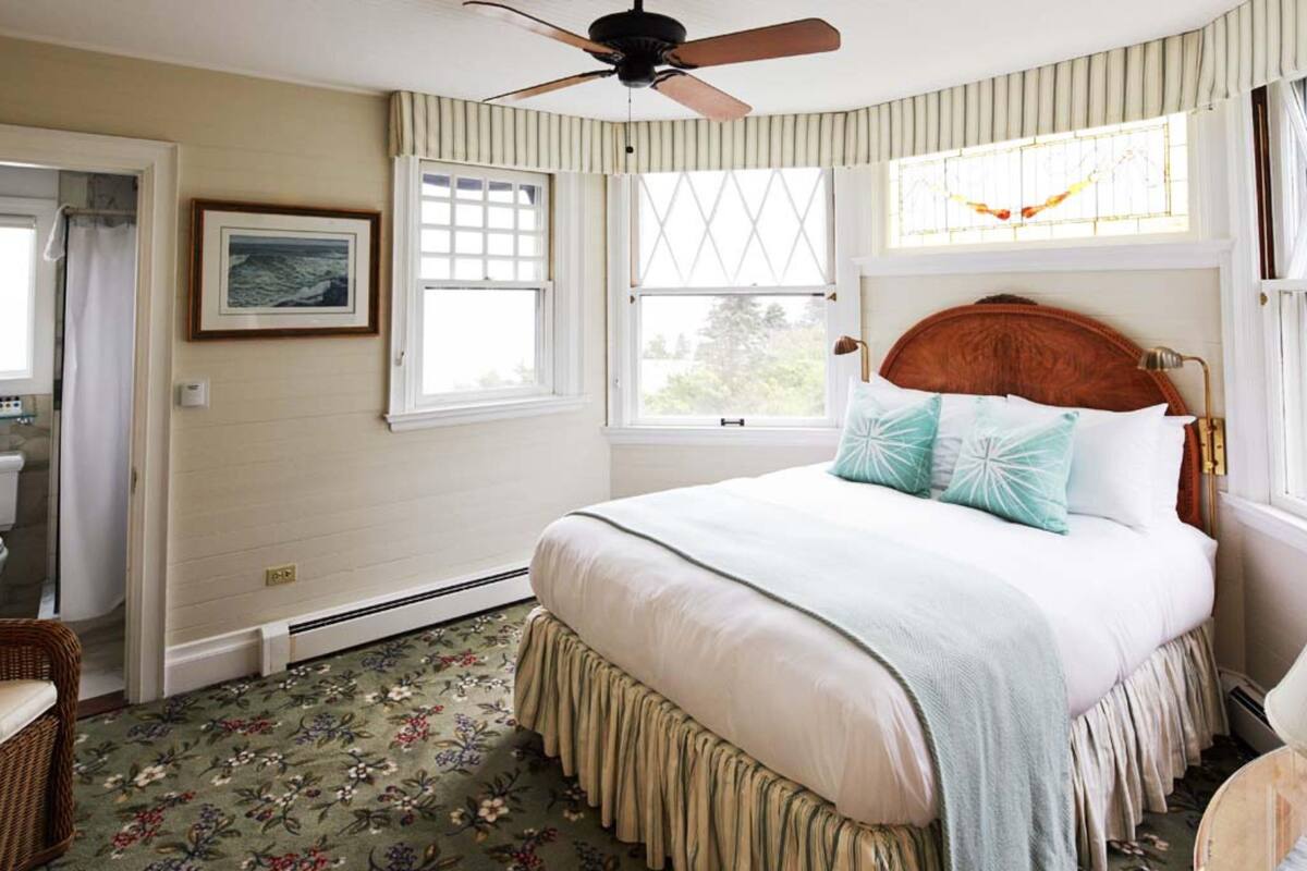 A bedroom with a bed, a ceiling fan, a window with a view, and a floral-patterned carpet.