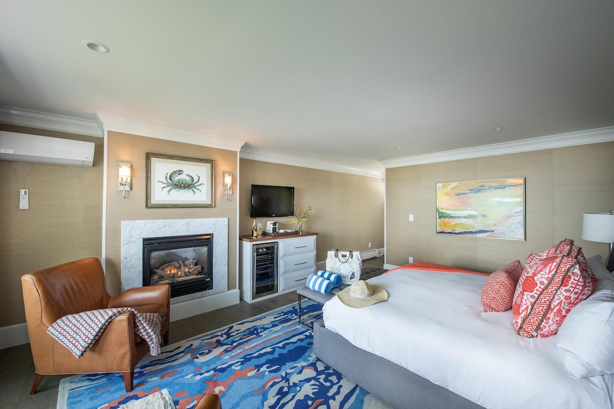 A hotel room with a fireplace, a bed with red and white pillows, a brown leather armchair, and a blue patterned rug.