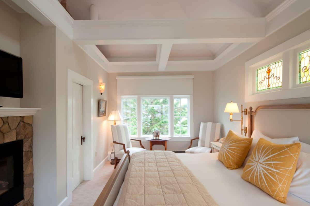 A bedroom with a large bed, a fireplace, a dining table, and a view of greenery outside.