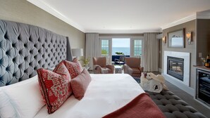 Una habitación de hotel con una cama grande, chimenea, vista al mar y un rincón de descanso con sillones.