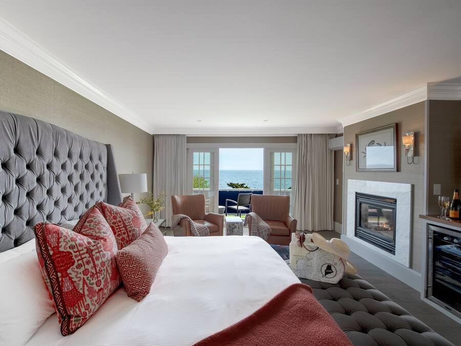 Una habitación de hotel con una cama grande, chimenea, vista al mar y un rincón de descanso con sillones.