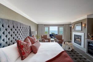 Una habitación de hotel con una cama grande, chimenea, vista al mar y un rincón de descanso con sillones.