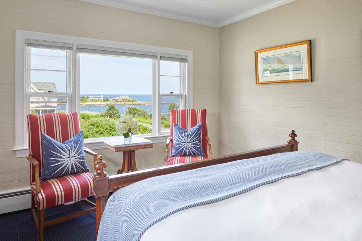 A bedroom with a large bed, two striped armchairs, a small table, and a window with a view of the ocean.