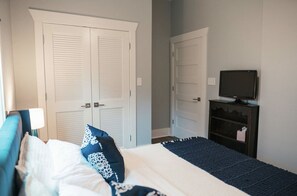 Un dormitorio con una cama, dos puertas blancas, un televisor sobre un mueble bajo y una mesita de noche.