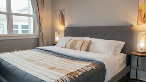 Een net opgemaakt bed met een grijs hoofdeinde, witte lakens en een gestreept plaid.