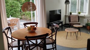 A dining area with a round table, chairs, and a fruit bowl. There is a television and a sofa in the background. - Beautiful detached house in an oasis of calm, close to beach and sea. (Koudekerke)