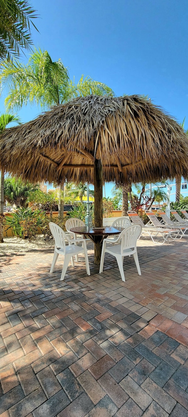 A thatched umbrella table with white plastic chairs, set on a paved patio with palm trees in the background.