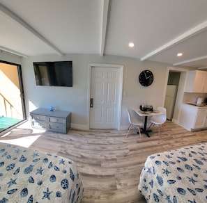 Un dormitorio con dos camas, televisión, puerta, reloj y un pequeño comedor con mesa y sillas.
