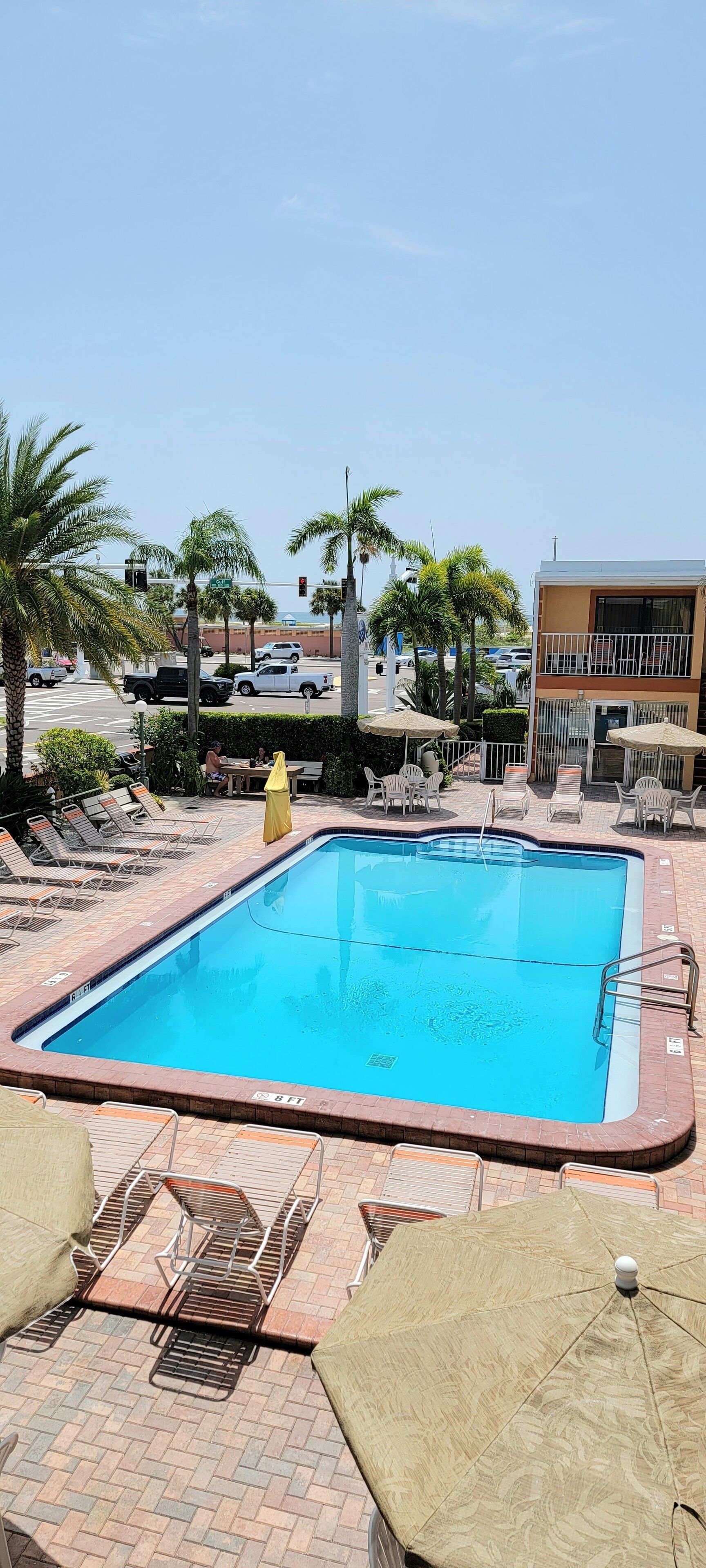 A swimming pool with lounge chairs and umbrellas, surrounded by palm trees and a building with a balcony.